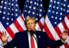 Man speaking on stage with American flags behind