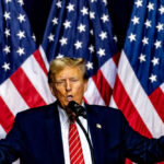 Man speaking on stage with American flags behind