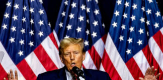 Man speaking on stage with American flags behind