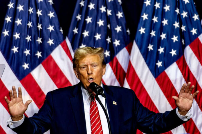 Man speaking on stage with American flags behind