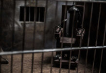An electric chair inside a dimly lit prison cell with bars in the foreground