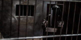 An electric chair inside a dimly lit prison cell with bars in the foreground