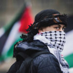 Person wearing keffiyeh and jacket with flag background