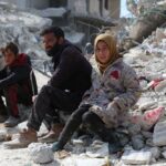 A man, a boy, and a girl sitting on rubble in a devastated area