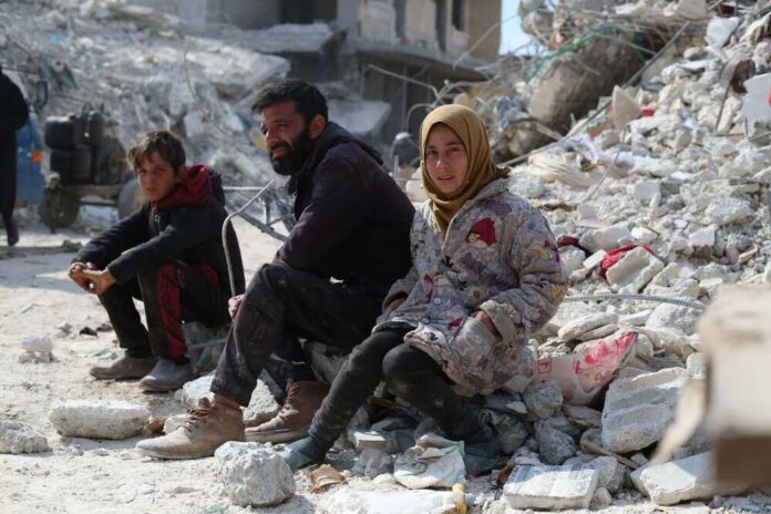 A man, a boy, and a girl sitting on rubble in a devastated area