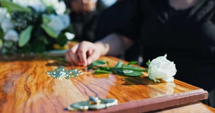 A hand placing a white rose on a wooden casket during a funeral service