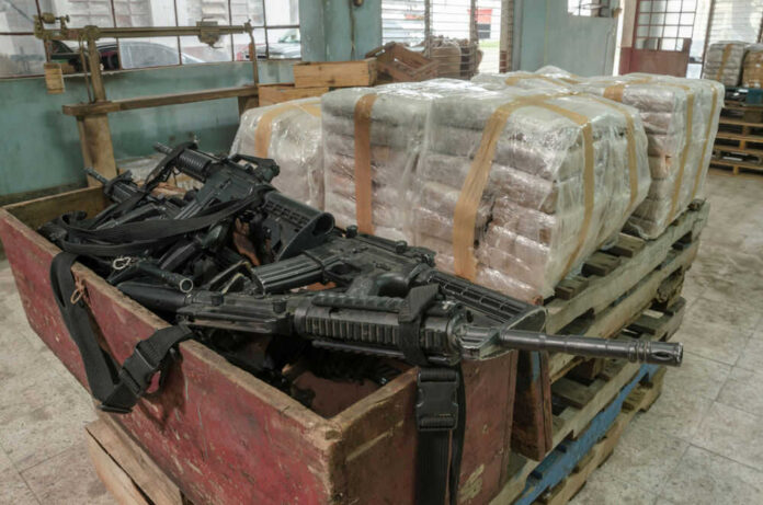 Pallets of wrapped packages beside a crate of firearms